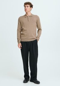 Polo de punto marrón de manga larga con cuello y tapeta de tres botones, combinado con pantalones negros con pliegues y zapatos negros.