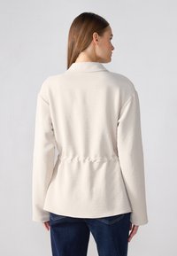 Lichtbeige jas met een relaxte pasvorm, kraag en een geraffineerde taille. Zacht, gestructureerd materiaal met lange mouwen en een ronde zoom.