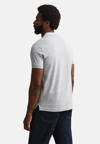 Man with curly hair and beard wearing a light gray short-sleeve polo shirt and dark blue jeans, facing left on a white background.