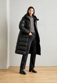 Schwarze Daunenjacke mit hohem Kragen und gestepptem Muster. Getragen über einem gestreiften Oberteil und kombiniert mit schwarzen Jeans und Stiefeletten mit Absatz.