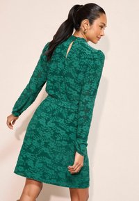 Zelen�é krajkové šaty s květinovým vzorem, dlouhými rukávy, páskem v pase a detailem ve tvaru klíčové dírky vzadu. Texturovaná látka s motivem listů.