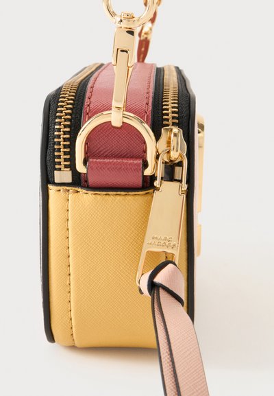 Πλάγια όψη μιας μικρής τσάντας Marc Jacobs με χρυσά φερμουάρ, κίτρινες και κόκκινες δερμάτινες επιφάνειες και ροζ-μαύρο ιμάντα καρπού.
