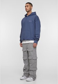 Mørkeblå oversized hettegenser med kengurulomme og hvit lagdelt crew neck. Grå cargo-bukser med flere lommer og en avslappet passform. Hvite sneakers.