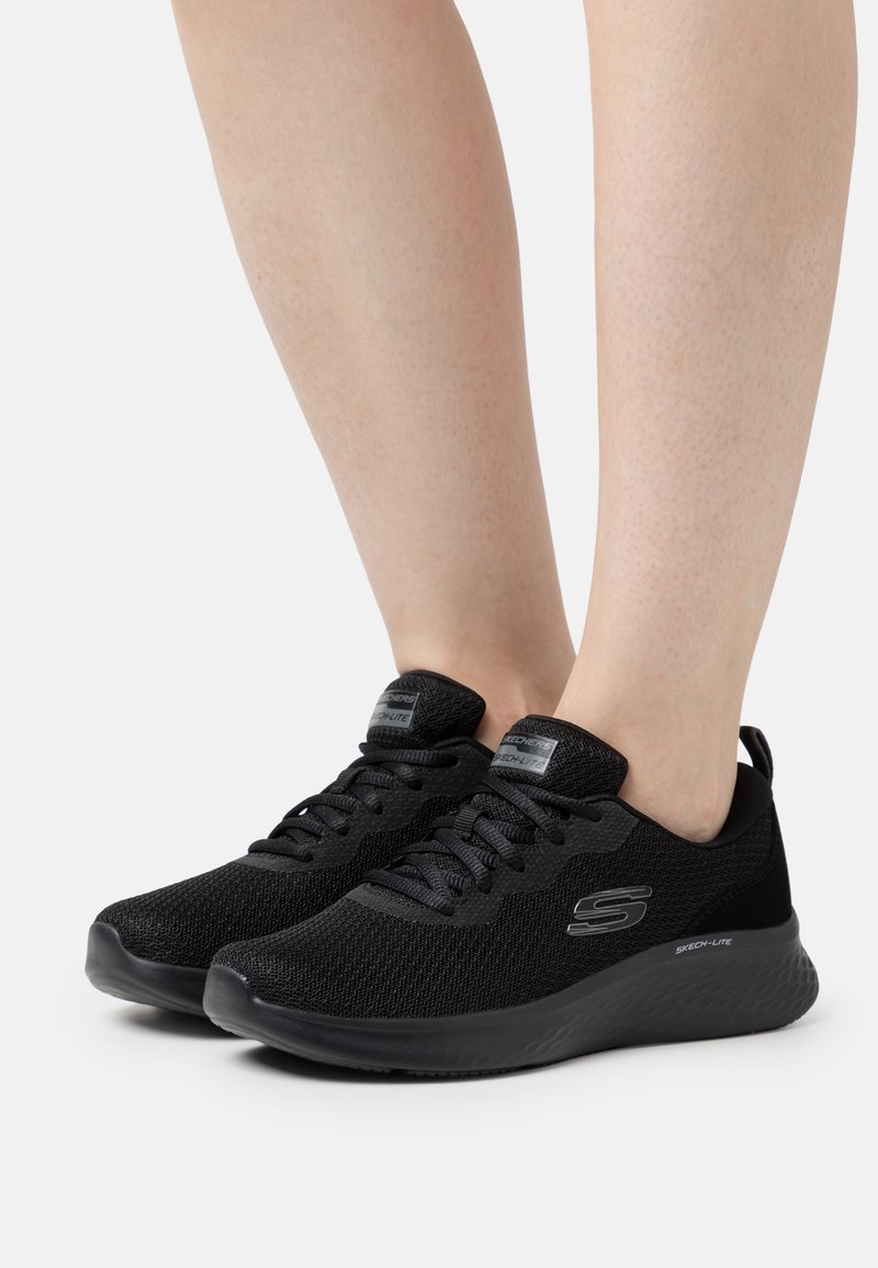 Jambes portant des baskets noires Skechers à lacets avec tige en mesh et semelle épaisse amortissante sur fond blanc.