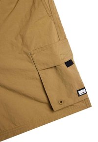 Khaki cargo shorts dengan kain berpola, dilengkapi saku flap, tab webbing hitam, dan label kecil bermerek dekat bagian bawah.