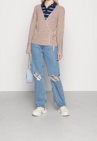 Beige omslagströja med långa ärmar och knyt detaljer, lagd över en marinblå randig polo. Ljusblå slitna jeans, vita chunky sneakers och en liten blå handväska.