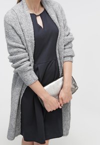 Cárdigan gris texturizado con mangas largas, llevado sobre un vestido oscuro. Sosteniendo un bolso metálico plateado. Diseño simple y tejido suave.