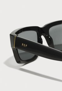 Gafas de sol negras con un marco grueso, lentes grises y el logo dorado de RSF en la patilla. Material plástico suave con un acabado brillante.