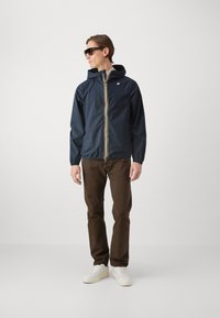 Marineblauwe waterdichte jas met een capuchon, voorzien van een gele rits, gecombineerd met bruine broeken en witte sneakers, staande tegen een neutrale achtergrond.