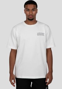 Bílý bavlněný tričko s krátkými rukávy, kulatým výstřihem a potištěným grafickým textem "WRONG FRIENDS" černou barvou na levé straně hrudi.