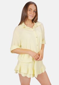 Ensemble de loungewear en coton jaune clair, comprenant une chemise à col, à manches courtes et à boutons, ainsi que des shorts à ourlet volanté, avec un tissu doux et texturé.
