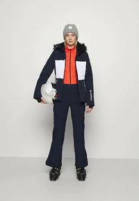 Σαπούνι σκι που περιλαμβάνει μια ναυτική ζακέτα με λευκές πάνελ, μια πορτοκαλί εσωτερική στρώση, ναυτικά παντελόνια και ένα γκρίζο καπέλο. Μαύρη μπότες σκι και κράνος συμπεριλαμβάνονται.