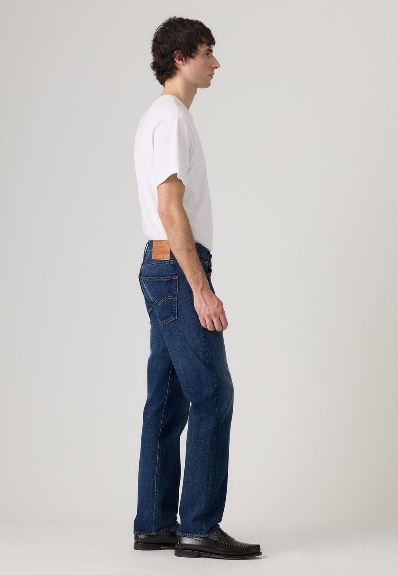 Young man standing in profile wearing a white short-sleeve shirt, dark blue jeans, and black leather shoes against a plain background.
