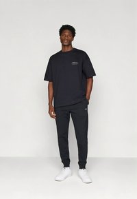 Fekete pamut póló rövid ujjakkal és kis fehér mintával, fekete nadrággal és fehér sportcipővel párosítva, laza, hétköznapi összeállítást alkotva.