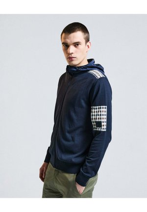 Giovane uomo che indossa una felpa con zip blu navy con toppa a quadri sulla spalla e sul braccio, e pantaloni verde oliva, in piedi davanti a uno sfondo uniforme.