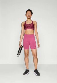 Atletische vrouw die staat in een roze sportbeha en shorts, een zwarte waterfles vasthoudend, gekleed in zwart-witte hardloopschoenen.