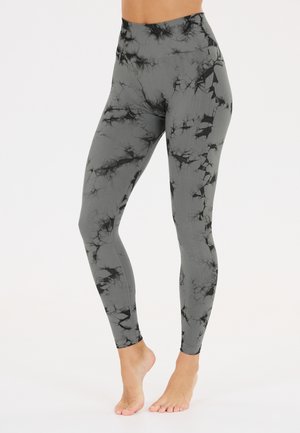 Kvinde-ben iført grå leggings med høj talje og sort batikmønster, stående barfodet på en hvid flade.