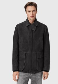 Homme portant une veste en daim noire avec des poches �à rabat et un col à sangle boutonné, associée à un jean gris clair, sur un fond uni.