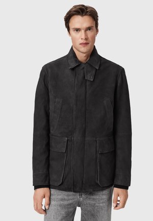 Man wearing a black suede jacket with flap pockets and a buttoned strap collar, paired with light gray jeans, against a plain background.