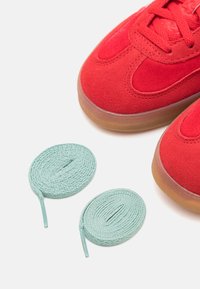 Baskets en daim rouge avec une semelle en caoutchouc, présentant des accents texturés et deux lacets en cuir roulés de couleur menthe à côté.