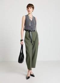 Graue, kurze Weste mit vorderen Knöpfen, hoch taillierte grüne Hose, schwarzer Gürtel und schwarze spitze Ballerinas. Trägt eine schwarze Umhängetasche.