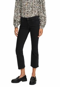 Persona con jeans negros recortados, blusa de manga larga con estampado de leopardo y mocasines de cuero negro, de pie contra un fondo blanco.