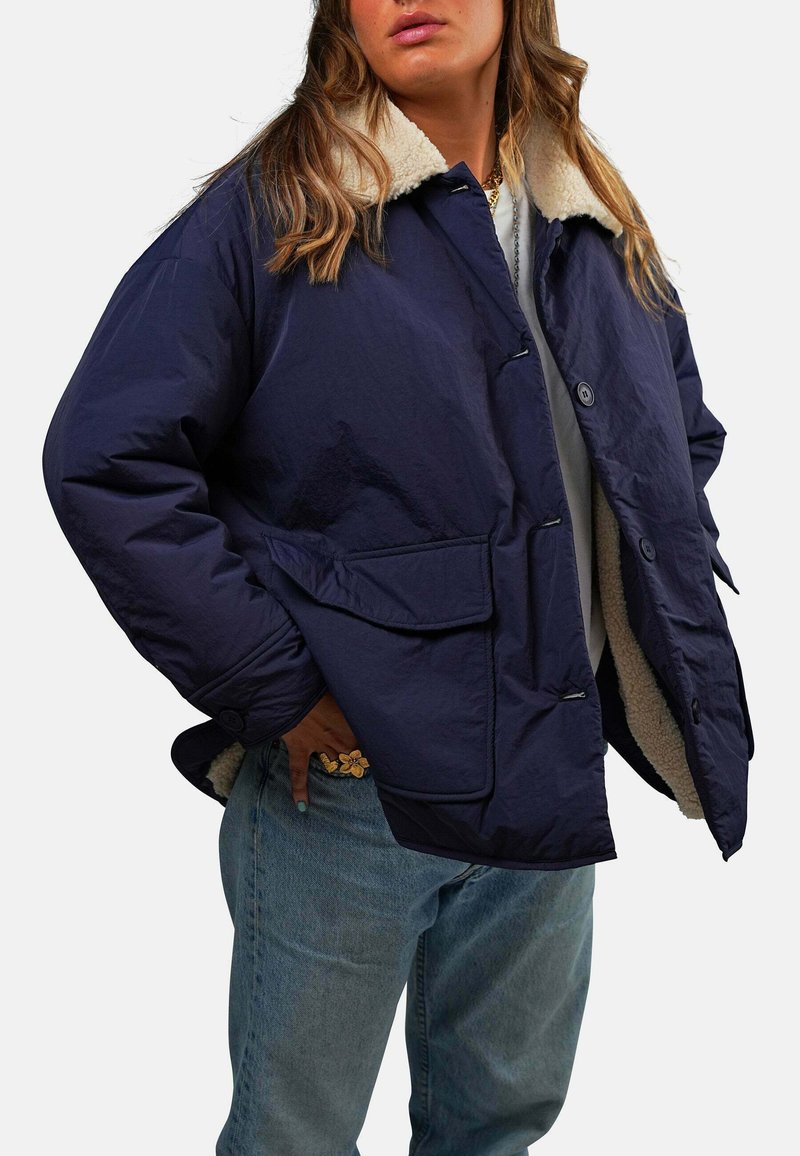 Veste bleu marine avec un col en polaire crème doux, dotée d'une fermeture à boutons et de deux poches avant. Portée par-dessus une chemise grise et un jean.