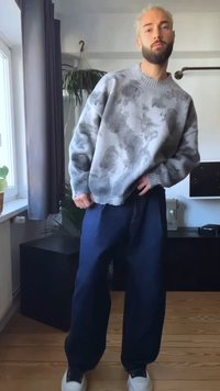 Sweater cinza com textura abstrata, punhos canelados, combinado com calças largas azul-escuro e ténis brancos, em pé sobre um chão de madeira.