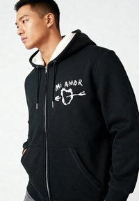 Zwarte hoodie met ritssluiting en een met schapenvacht gevoerde capuchon. Bevat witte grafische tekst "Mi Amor" en een pijlontwerp. Voorzakken en een zilveren rits.