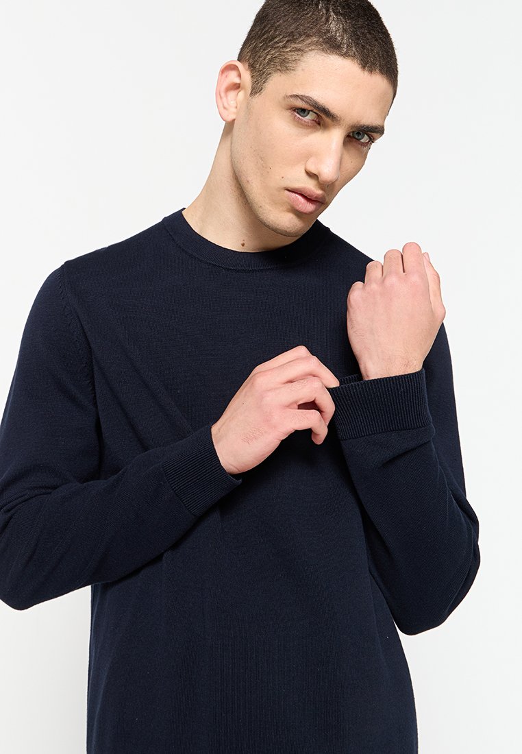 Pull en jersey bleu marine foncé en tissu doux et texturé. Présente des poignets côtelés et une silhouette ajustée, porté avec des mains ajustant la manche.