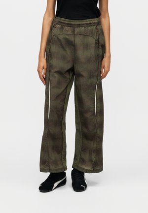 Personne portant un pantalon ample à carreaux vert olive avec des fermetures éclair latérales et des baskets noires Puma, debout devant un fond uni.