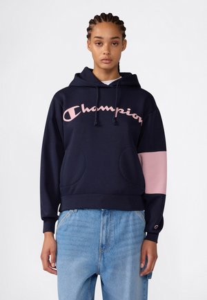 Junge Person mit geflochtenem Haar, die einen marineblauen Hoodie mit pinkem Champion-Logo und pinkem Ärmelpaneel sowie hellblaue Jeans trägt.