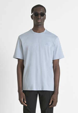 T-shirt azzurro chiaro a maniche corte realizzato in morbido cotone, con collo a girocollo e un piccolo dettaglio con logo sul lato sinistro del petto. Indossato con pantaloni neri.