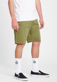 Mand iført olivengrønne shorts, hvide sokker med sorte logoer og sorte slip-on sneakers, stående foran en ensfarvet hvid baggrund.