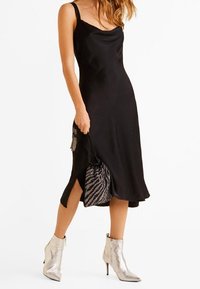 Robe nuisette en satin noir avec un décolleté drapé, fente latérale, associée à des bottines argentées et un sac rayé noir et blanc.
