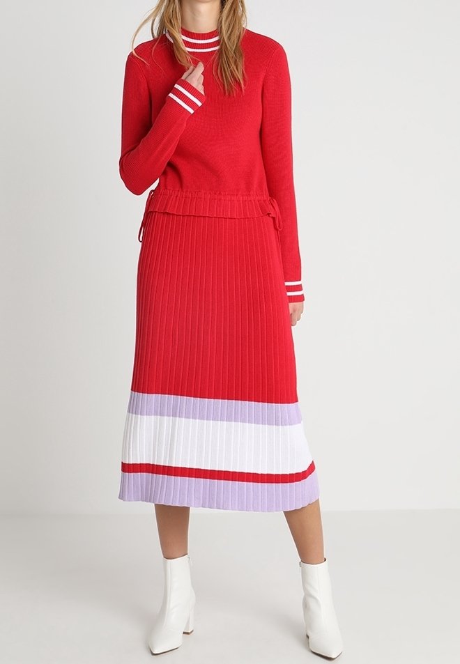 Femme portant une robe rouge à manches longues avec des bandes blanches et lavande à l'ourlet et des bottines blanches, debout sur un fond uni.