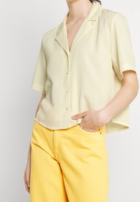 Blouse jaune clair à manches courtes avec col et devant boutonné, confectionnée dans un tissu lisse. Associée à un pantalon jaune.