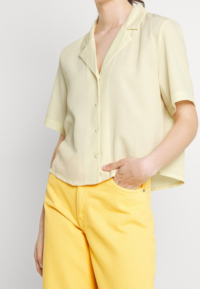 Blouse jaune clair à manches courtes avec col et devant boutonné, confectionnée dans un tissu lisse. Associée à un pantalon jaune.