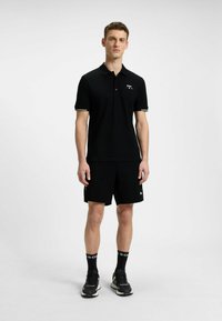 Czarna koszulka polo z kołnierzykiem i trzema guzikami, z złotymi akcentami. W zestawie z czarnymi szortami i czarnymi butami sportowymi, całość uzupełnia stylizację.