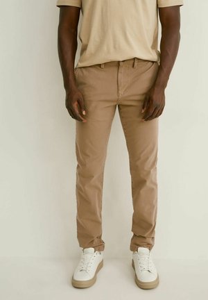 Pantaloni beige slim fit indossati con una camicia beige abbinata e sneakers bianche su una persona in piedi davanti a uno sfondo semplice.