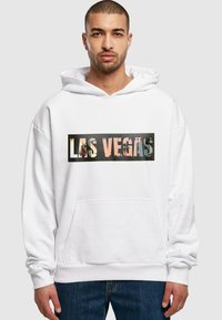 Baltas džemperis su priekiniu kišeniu ir ryškiu "LAS VEGAS" grafiniu motyvu juodame stačiakampyje. Audinys atrodo minkštas ir turi laisvą siluetą.