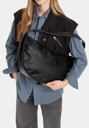 Donna con camicia grigia e gilet nero tiene una grande borsa a tracolla in pelle nera di G-Star attraversata sul torso.