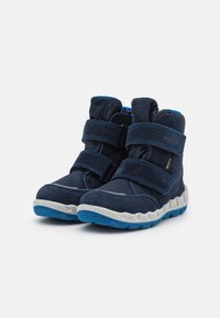 Botas azul marino con dos correas de velcro, parte superior de tejido texturizado, collar acolchado y una suela de goma blanca ondulada con acentos azules.