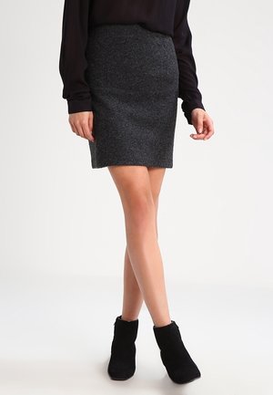 Kvinde iført en mørkegrå nederdel, sort langærmet top og sorte ankelstøvler, stående med benene krydset mod en ensfarvet baggrund.