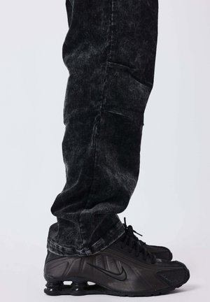 Persona che indossa pantaloni in velluto cord e sneakers Nike nere con ammortizzatori a molla visibili, in piedi su uno sfondo chiaro.