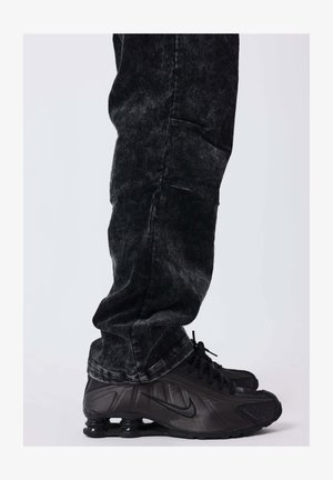 Persoon in zwarte corduroybroek en zwarte Nike sneakers met zichtbare veerachtige zolen, staand op een lichte achtergrond.