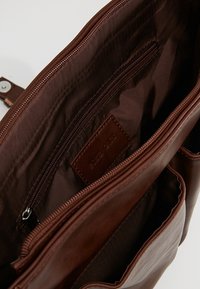 Interiér hnědé kožené tašky s tmavým podšívkou, kapsou na zip a značkovou koženou visačkou. Hladká textura a strukturovaný design.