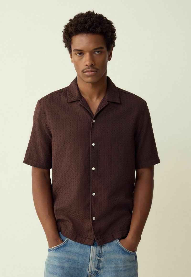 Short-sleeve brown shirt with a textured pattern, button-down front, and a spread collar, paired with light blue jeans.