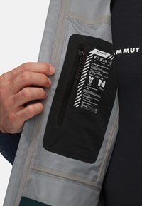 Poche zippée noire sur une veste grise, avec des instructions de sécurité en graphiques blancs. Tissu lisse avec un léger éclat.