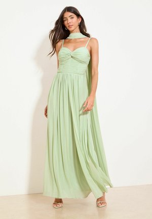 Žena nosiaca prúdiace svetlozelené šaty bez rukávov so shromáždeným živôtikom a zodpovedajúcim šálom previazaným okolo krku a plecí.